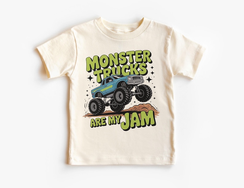Puede incluir: Camiseta color crema con un gr&aacute;fico de cami&oacute;n monstruo azul y el texto "MONSTER TRUCKS ARE MY JAM" en verde y negro. El dise&ntilde;o incluye estrellas y un mont&iacute;culo de tierra marr&oacute;n.