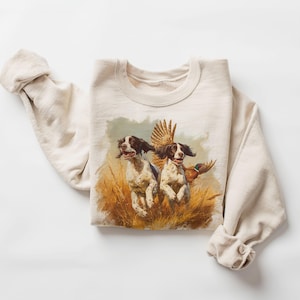 Puede incluir: Sudadera color crema con una ilustración de estilo vintage de dos perros corriendo por la hierba alta, con un faisán en vuelo. Los perros son blancos y negros, y el faisán tiene plumas marrones y naranjas.