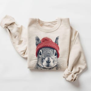 May include: Cream-colored sweatshirt featuring a detailed illustration of a squirrel wearing a red knit beanie. The squirrel's face is the focal point, with its whiskers and eyes clearly visible. The sleeves are rolled up, and the overall style is casual.