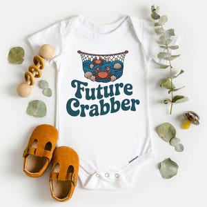 May include: White baby bodysuit with a crab and ocean graphic, and the text "Future Crabber" in teal. The bodysuit is surrounded by a wooden toy, baby shoes, and decorative leaves.