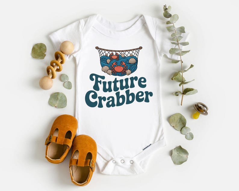May include: White baby bodysuit with a crab and ocean graphic, and the text "Future Crabber" in teal. The bodysuit is surrounded by a wooden toy, baby shoes, and decorative leaves.