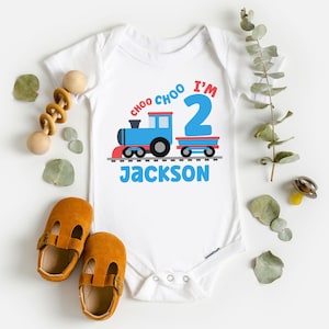 May include: White baby onesie with a blue train and red caboose. The train has the words "Choo Choo I'm 2" on it. The onesie is personalized with the name "Jackson".