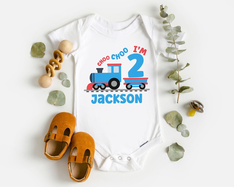 May include: White baby onesie with a blue train and red caboose. The train has the words "Choo Choo I'm 2" on it. The onesie is personalized with the name "Jackson".