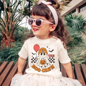May include: A child wearing a cream-colored t-shirt with a Halloween-themed graphic. The graphic features a ghost holding a balloon and a pumpkin, with the text "The Boo Crew". The child is also wearing pink sunglasses and a pink headband.