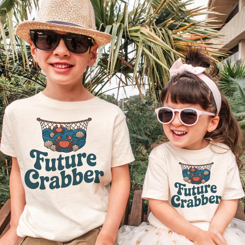 May include: Two children wearing cream-colored t-shirts with a graphic of a crab in a net, seashells, and the words "Future Crabber" in teal. One child wears a straw hat and sunglasses, the other wears a pink headband and sunglasses.