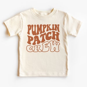 May include: Cream-colored t-shirt with the words "PUMPKIN PATCH CREW" in a retro, brown font. The short-sleeved shirt is made of a soft material, perfect for fall activities. Ideal for kids.