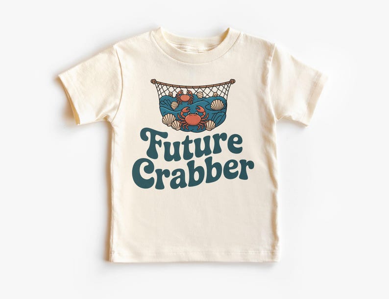 May include: Cream-colored t-shirt featuring a graphic of a crab net with crabs, seashells, and waves. The text "Future Crabber" is in teal, retro-style lettering. A fun, nautical-themed tee for kids.