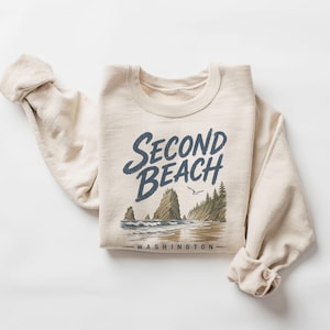May include: A cream-colored crewneck sweatshirt featuring a graphic print of a coastal landscape with rock formations, pine trees, and a bird. The text "SECOND BEACH" is printed in large dark blue letters above the scene, with "WASHINGTON" below.