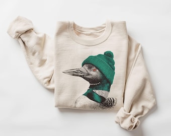 Puede incluir: Sudadera de color crema con una ilustración detallada de un colimbo luciendo un gorro de punto verde con un pompón. El colimbo tiene una bufanda a rayas verdes y blancas. Las mangas están dobladas. La sudadera está tendida sobre una superficie blanca.