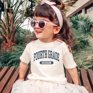 May include: A young child wearing a cream-colored t-shirt with the words "FOURTH GRADE" in navy blue, with "HUDSON" below. The child is also wearing pink sunglasses and a matching headband. The child is sitting on a wooden bench.