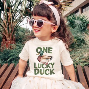 Puede incluir: Una ni&ntilde;a lleva una camiseta color crema con las palabras "ONE LUCKY DUCK" y una ilustraci&oacute;n de un pato. La ni&ntilde;a tambi&eacute;n lleva gafas de sol rosas y una diadema a juego. La ni&ntilde;a est&aacute; sentada en un banco de madera.