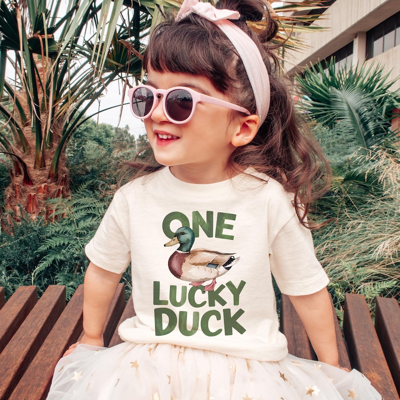 Puede incluir: Una ni&ntilde;a lleva una camiseta color crema con las palabras "ONE LUCKY DUCK" y una ilustraci&oacute;n de un pato. La ni&ntilde;a tambi&eacute;n lleva gafas de sol rosas y una diadema a juego. La ni&ntilde;a est&aacute; sentada en un banco de madera.