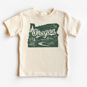 May include: A cream colored t-shirt with a green outline of the state of Oregon. The outline includes a mountain range, a river, trees, and a lighthouse. The word "Oregon" is written in a vintage style font.