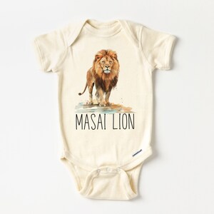 May include: A cream-colored baby onesie with a watercolor illustration of a lion standing in a puddle of water. The text "MASAI LION" is printed in black below the lion.