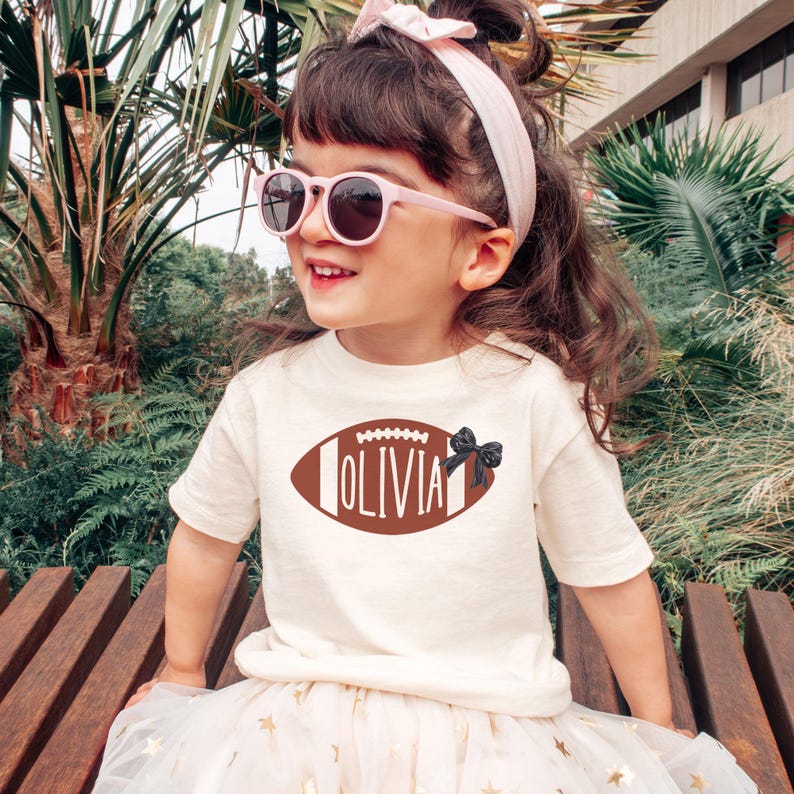 Puede incluir: Una ni&ntilde;a lleva una camiseta crema con un dise&ntilde;o de bal&oacute;n de f&uacute;tbol americano marr&oacute;n y el nombre "Olivia". La ni&ntilde;a tambi&eacute;n lleva gafas de sol rosas, una diadema rosa y una falda tut&uacute; blanca con estrellas doradas. La ni&ntilde;a est&aacute; sentada en un banco de madera.
