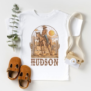 May include: White t-shirt with a graphic of a cowboy riding a horse in a desert landscape. The graphic includes a sunset, cacti, and the text "HUDSON".