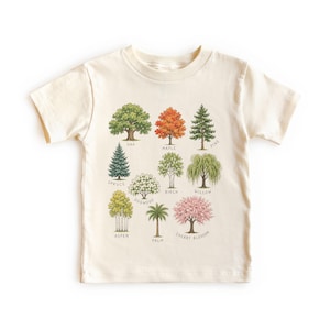May include: A cream-colored short-sleeve t-shirt featuring a graphic print of twelve distinct trees. The illustration includes labels for Oak, Maple, Pine, Spruce, Dogwood, Birch, Willow, Aspen, Palm, and Cherry Blossom in black text.