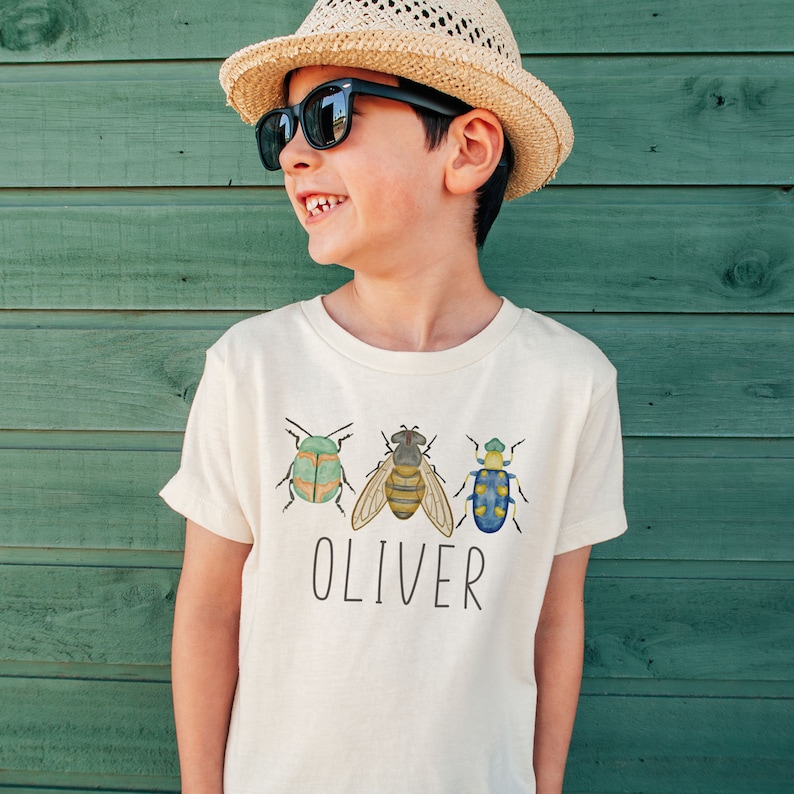 May include: A white t-shirt with a graphic of three insects, a green beetle, a bee, and a blue beetle. The text "OLIVER" is printed below the insects.