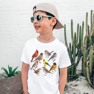 以下が含まれることがあります： さまざまな鳥のカラフルなイラストが描かれた白いTシャツ。デザインには、赤いカージナル、ルリビタキ、その他の鳥が含まれています。Tシャツは、サングラスと帽子をかぶった人が着用しています。