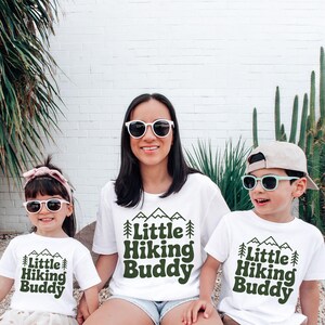 May include: Three people wearing white t-shirts with the text "Little Hiking Buddy" in green, along with mountain and tree graphics. They are also wearing sunglasses. The background features plants and a white brick wall.