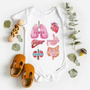 K&ouml;nnte beinhalten: Wei&szlig;er Baby-Strampler mit einer bunten Illustration der inneren Organe des menschlichen K&ouml;rpers, darunter Herz, Lunge, Leber, Magen, Darm und Nieren. Der Text 'LUNGS', 'HEART', 'LIVER', 'STOMACH', 'INTESTINES' und 'KIDNEYS' ist auf dem Strampler aufgedruckt.