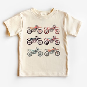 May include: A cream-colored t-shirt with a graphic of six vintage dirt bikes in different colors. The bikes are arranged in three rows of two.