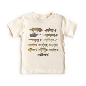 以下が含まれることがあります： クリーム色の半袖Tシャツで、様々な魚種が列になって並ぶ詳細なグラフィックプリントが施されています。各魚には、アラパイマ、アリゲーターガー、ベルーガチョウザメなど、小さな黒い文字で一般的な名前が付けられています。
