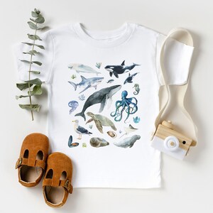 May include: White t-shirt with a watercolor design of various sea creatures, including a whale, octopus, sea turtle, and seahorse. The shirt is on a white background with a wooden toy camera and brown shoes.