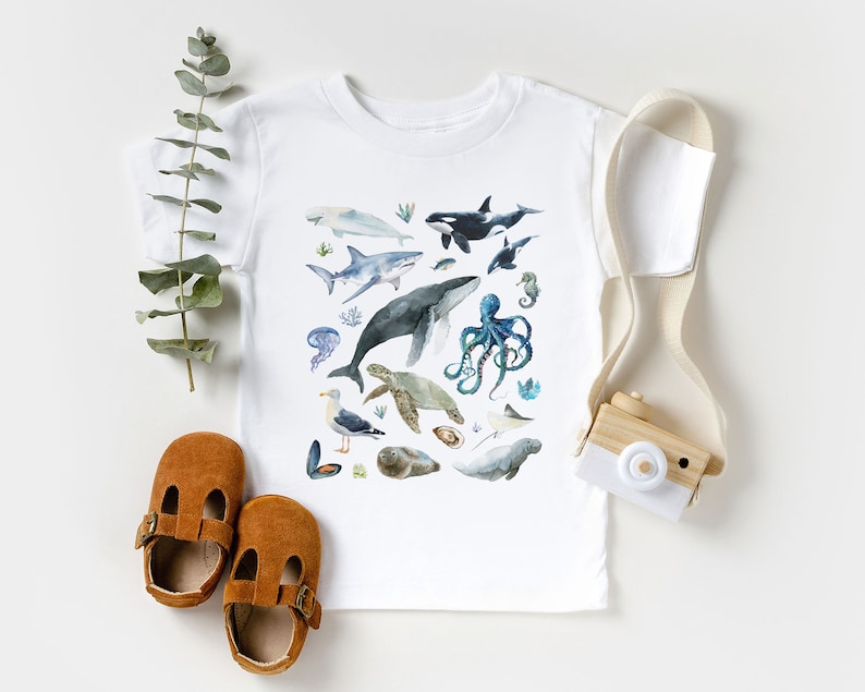 May include: White t-shirt with a watercolor design of various sea creatures, including a whale, octopus, sea turtle, and seahorse. The shirt is on a white background with a wooden toy camera and brown shoes.