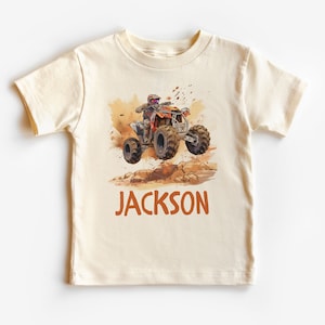 May include: A beige t-shirt with a watercolor illustration of a person riding an orange ATV on a dirt track. The text "JACKSON" is printed in orange below the image.