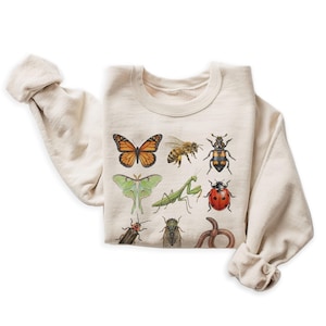 May include: Cream-colored sweatshirt featuring a collection of illustrated insects. The design includes a monarch butterfly, a bee, a ladybug, a praying mantis, and other bugs. The sweatshirt has a crew neck and long sleeves.