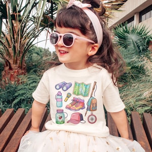 K&ouml;nnte beinhalten: Ein Kind tr&auml;gt ein cremefarbenes T-Shirt mit farbenfrohen Camping-Motiven, darunter ein Fernglas, eine Karte und Wanderschuhe. Das Kind tr&auml;gt au&szlig;erdem eine rosa Sonnenbrille, ein passendes Stirnband und einen wei&szlig;en Rock.