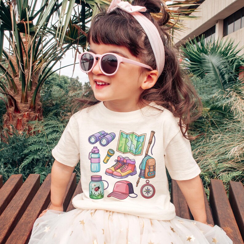 K&ouml;nnte beinhalten: Ein Kind tr&auml;gt ein cremefarbenes T-Shirt mit farbenfrohen Camping-Motiven, darunter ein Fernglas, eine Karte und Wanderschuhe. Das Kind tr&auml;gt au&szlig;erdem eine rosa Sonnenbrille, ein passendes Stirnband und einen wei&szlig;en Rock.