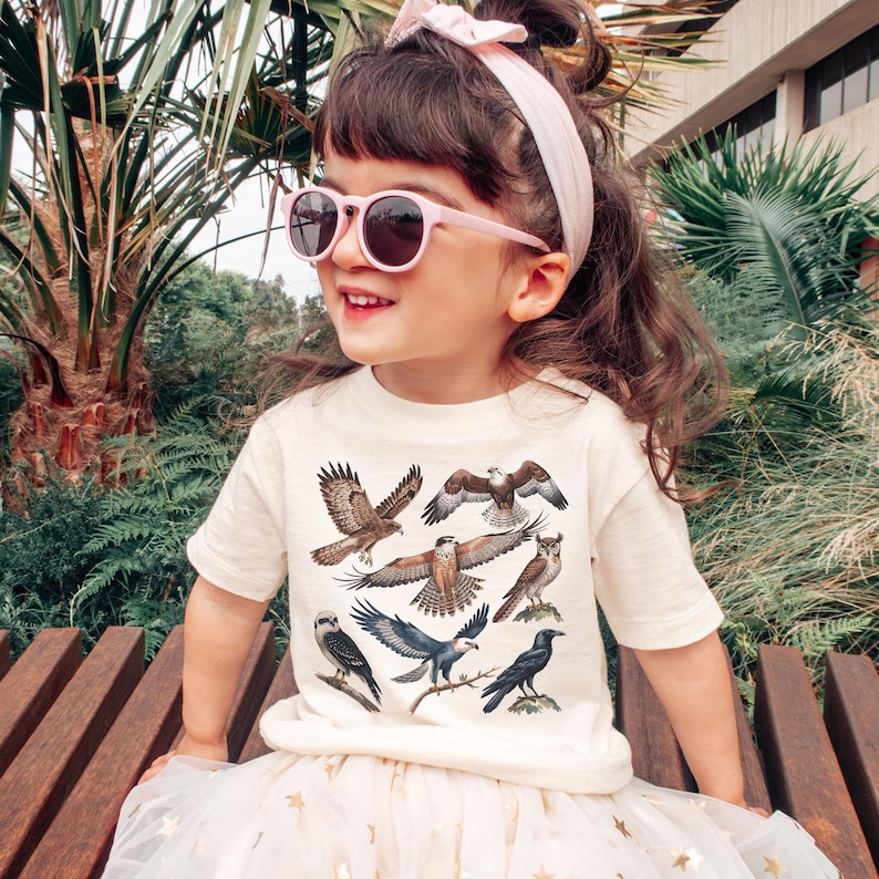 K&ouml;nnte beinhalten: Ein Kind tr&auml;gt ein cremefarbenes T-Shirt mit Illustrationen verschiedener Greifv&ouml;gel. Das Kind tr&auml;gt auch eine rosa Sonnenbrille und ein passendes Stirnband. Das Kind sitzt auf einer Holzbank.