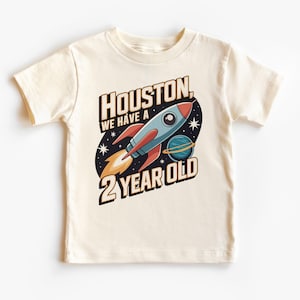 Puede incluir: Una camiseta blanca con un diseño de cohete y el texto "Houston, we have a 2 year old".