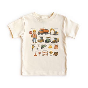 May include: A cream-colored short-sleeve t-shirt featuring a graphic print of construction equipment and a worker. The illustration includes a dump truck, excavator, road roller, asphalt paver, backhoe, jackhammer, shovel, wheelbarrow, and safety gear.