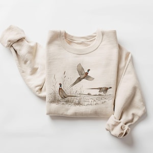 Op de afbeelding: Een crèmekleurige sweater met ronde hals en een gedetailleerde grafische illustratie van drie fazanten in een veld. Eén fazant staat in het hoge gras, terwijl twee andere vliegend zijn afgebeeld tegen een natuurlijke landschapsachtergrond.