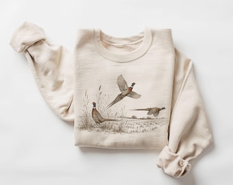 May include: A cream-colored crewneck sweatshirt featuring a detailed graphic illustration of three pheasants in a field. One pheasant stands in tall grass while two others are depicted in flight against a natural, muted landscape background.