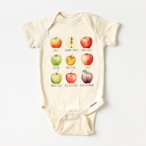 May include: A beige baby onesie with a graphic of nine different types of apples, each with its name printed below. The apples are illustrated in a watercolor style. The apple varieties are Gala, Granny Smith, Honeycrisp, Crispin, Pink Pearl, Fuji, Ginger Gold, Red Delicious, and Black Diamond.