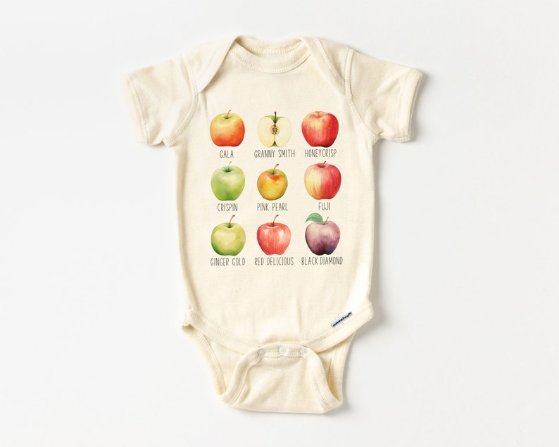 May include: A beige baby onesie with a graphic of nine different types of apples, each with its name printed below. The apples are illustrated in a watercolor style. The apple varieties are Gala, Granny Smith, Honeycrisp, Crispin, Pink Pearl, Fuji, Ginger Gold, Red Delicious, and Black Diamond.