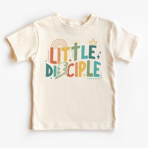 Könnte beinhalten: Ein cremefarbenes T-Shirt mit kurzen Ärmeln und dem Text "LITTLE DISCIPLE" in bunten Buchstaben mit einem Regenbogen, einem Kreuz, einem Blitz und einer Krone.