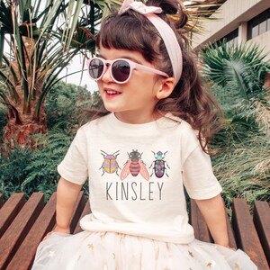 以下が含まれることがあります： 3匹のカラフルな虫と「Kinsley」という名前がプリントされた白いTシャツ。ピンクのサングラスとピンクのヘッドバンドをつけた子供が着用しています。