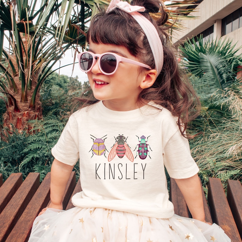 以下が含まれることがあります： 3匹のカラフルな虫と「Kinsley」という名前がプリントされた白いTシャツ。ピンクのサングラスとピンクのヘッドバンドをつけた子供が着用しています。