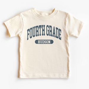 May include: Cream-colored short-sleeved t-shirt with the words "FOURTH GRADE" in navy blue arched across the chest. Below the text is the name "HUDSON" in a navy blue oval. The shirt is made of a soft material.