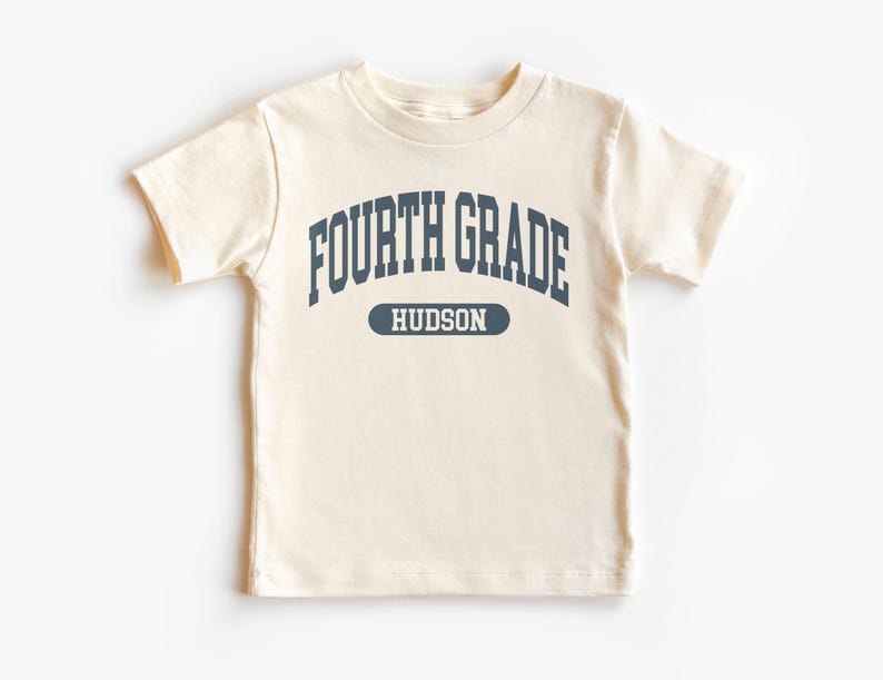 May include: Cream-colored short-sleeved t-shirt with the words "FOURTH GRADE" in navy blue arched across the chest. Below the text is the name "HUDSON" in a navy blue oval. The shirt is made of a soft material.