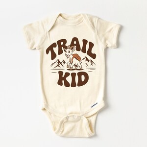 Peut inclure: Body de b&eacute;b&eacute; cr&egrave;me avec l'inscription "TRAIL KID" en marron, dans une typographie r&eacute;tro. Une ch&egrave;vre de dessin anim&eacute; avec un sac &agrave; dos est centr&eacute;e entre les mots, avec des graphiques de montagnes au-dessus d'une ligne horizontale. Le body a des manches courtes et des fermetures &agrave; pression.