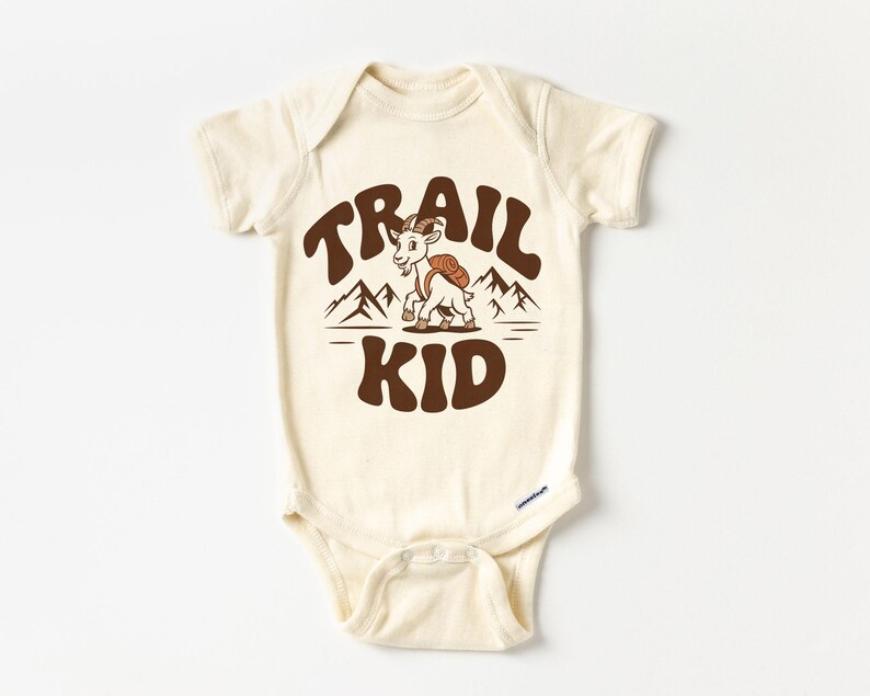 Peut inclure: Body de b&eacute;b&eacute; cr&egrave;me avec l'inscription "TRAIL KID" en marron, dans une typographie r&eacute;tro. Une ch&egrave;vre de dessin anim&eacute; avec un sac &agrave; dos est centr&eacute;e entre les mots, avec des graphiques de montagnes au-dessus d'une ligne horizontale. Le body a des manches courtes et des fermetures &agrave; pression.