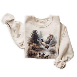 May include: A cream-colored sweatshirt with a graphic print of ducks flying over a lake in a forest setting.
