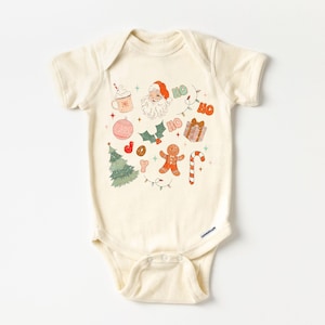 Puede incluir: Body de beb&eacute; color crema con un dise&ntilde;o navide&ntilde;o festivo. El dise&ntilde;o incluye a Pap&aacute; Noel, un &aacute;rbol de Navidad, un hombre de jengibre, un bast&oacute;n de caramelo, adornos y la palabra "HO" en naranja. Tambi&eacute;n se muestra una taza de chocolate caliente y la palabra "JOY".