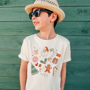 Puede incluir: Una camiseta color crema con un dise&ntilde;o navide&ntilde;o festivo. El gr&aacute;fico incluye a Pap&aacute; Noel, un hombre de jengibre, un &aacute;rbol de Navidad y la palabra "HO". La camiseta la lleva un ni&ntilde;o con gafas de sol y un sombrero de paja.